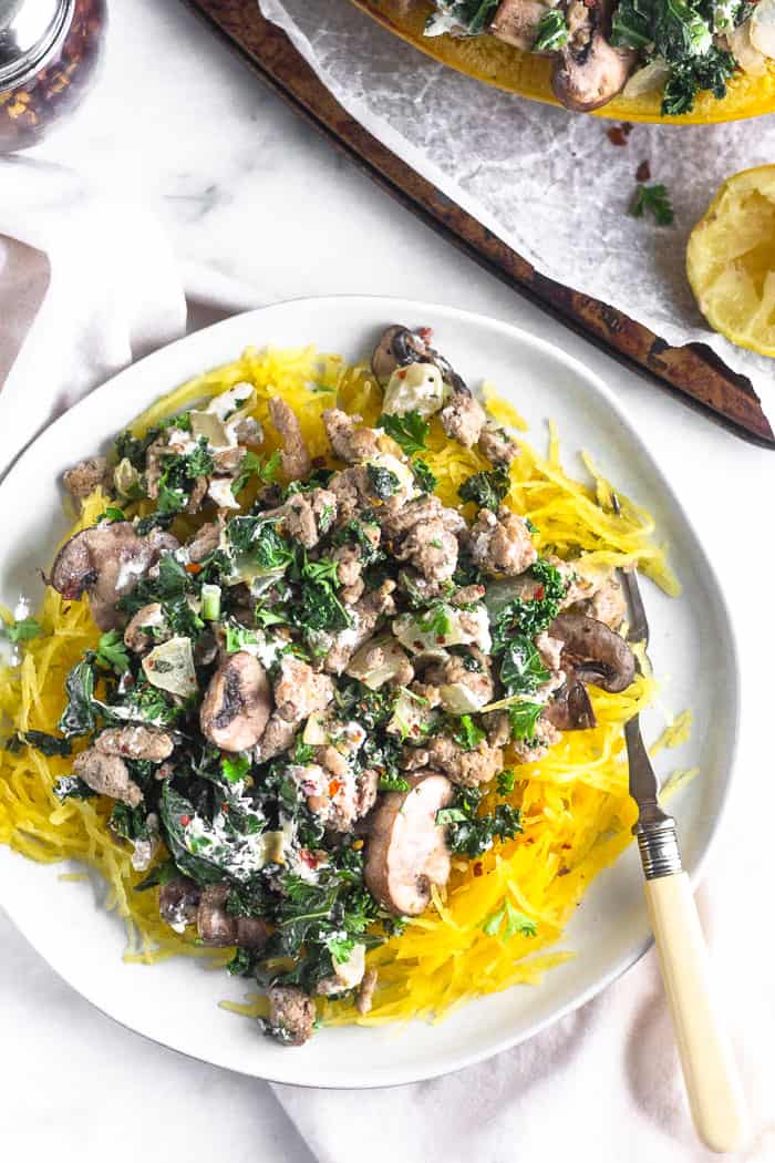 A white plate filled with spaghetti squash with ground turkey, kale, mushrooms, and goat cheese. A fork is resting on the plate and the plate is on top of a white linen.