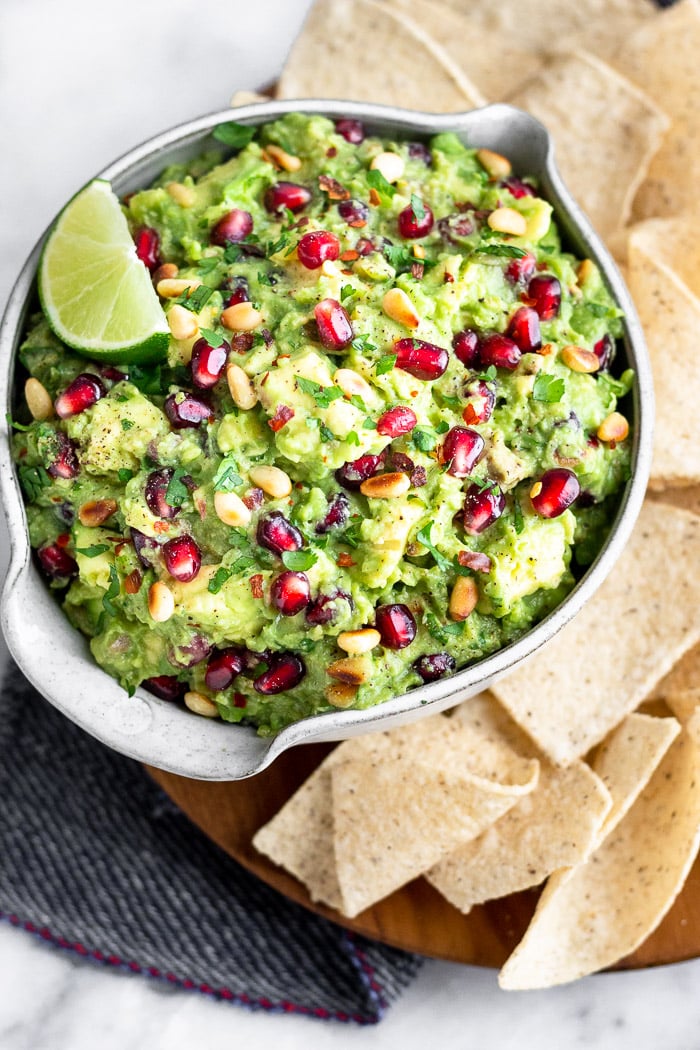 A wooden plate with a bowl filled with pomegranate guacamole garnished with a lime wedge. On the plate is a bunch of chips surrounding the guacamole.