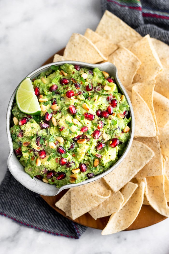 A bowl filled with pomegranate guacamole with chips surrounding it on a wooden plate. The plate is on a blue linen.