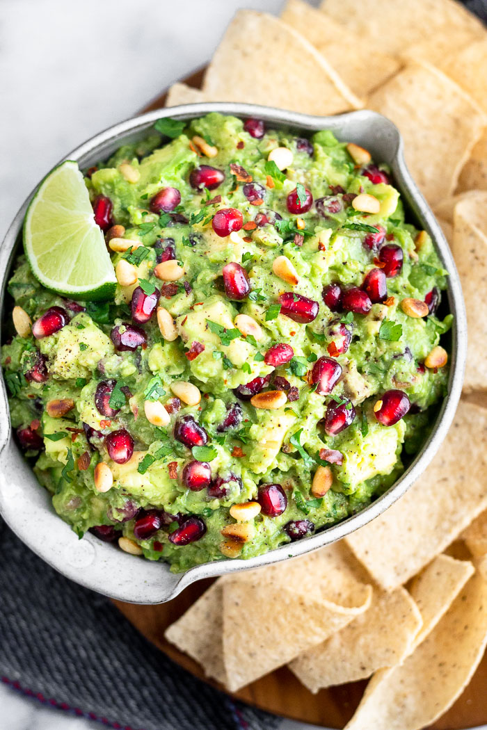 Close up of a bowl filled with pomegranate guacamole topped with a lime wedge. It is sitting on a plate that is filled with tortilla chips.