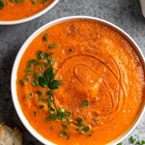 A bowl of roasted red pepper and squash soup stopped with chopped parsley and pepper. Around it is 2 pieces of bread, another bowl of soup, and a tan linen.