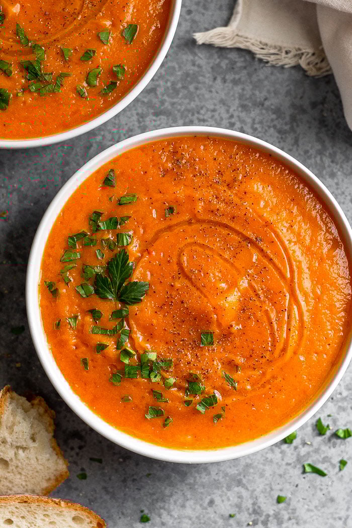 A bowl of roasted red pepper and squash soup stopped with chopped parsley and pepper. Around it is 2 pieces of bread, another bowl of soup, and a tan linen.