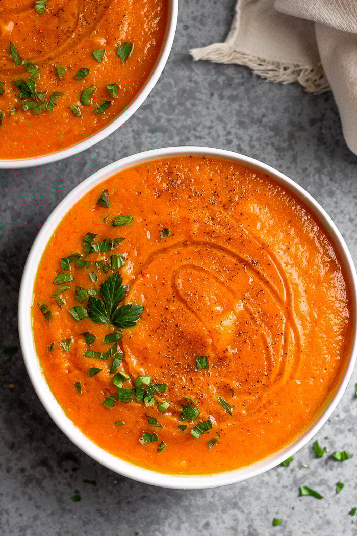 Bowl of acorn squash soup topped with chopped parsley and black pepper. More chopped parsley is around it, along with another bowl of soup and a tan linen.