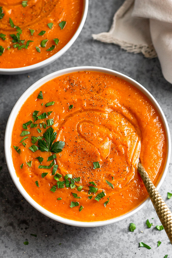 Bowl of vegan roasted red pepper and acorn squash soup topped with fresh parsley with a spoon in the bowl. It is surrounded by more parsley, another bowl, and a tan linen.