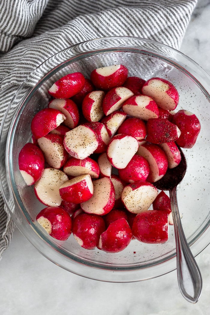 A bowl of chopped up radishes mixed with oil and spices. A spoon is coming out of the bowl and a stripped towel is next to it.