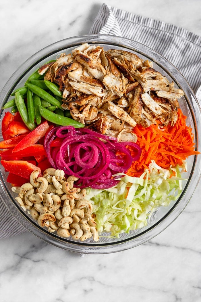 A large glass bowl filled with shredded cabbage, sliced red peppers, sugar snap peas, pulled chicken, shredded carrots, pickled onions, and cashews.