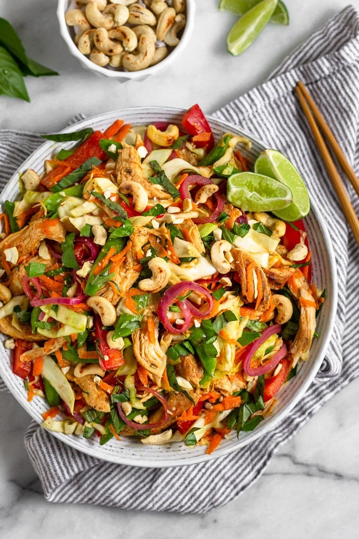 Overhead view of a bowl of chicken cabbage salad topped with fresh basil and garnished with lime wedges. Next to it is chopsticks, a bowl of cashews, lime wedges, and fresh Thai basil.