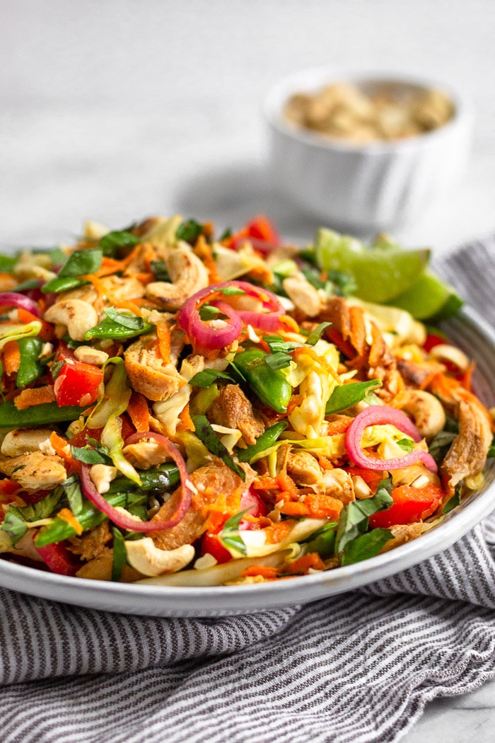 White shallow bowl filled with healthy chicken cabbage salad topped with fresh basil on sitting on a striped towel. A bowl of cashews is behind it.