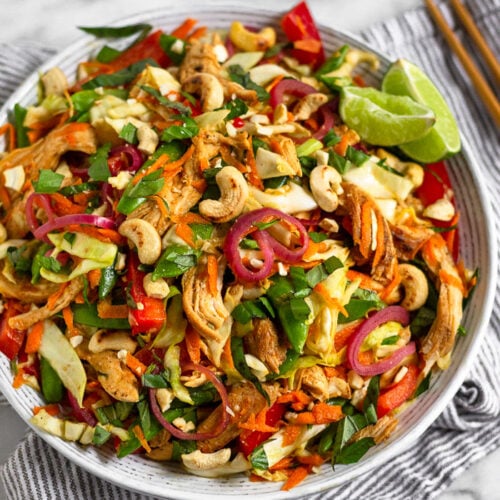 Vietnamese Chicken Cabbage Salad in a white bowl on a blue and white striped towel. Around it are chopsticks, a bowl of cashews, and lime wedges.