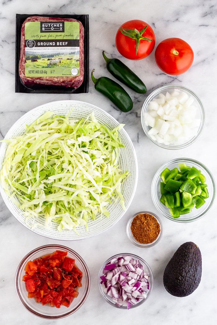 White counter filled with everything to make taco spaghetti squash boats. Ground beef, tomatoes, jalapeños, diced onion, diced pepper, avocado, taco seasoning, canned tomatoes, and shredded cabbage.