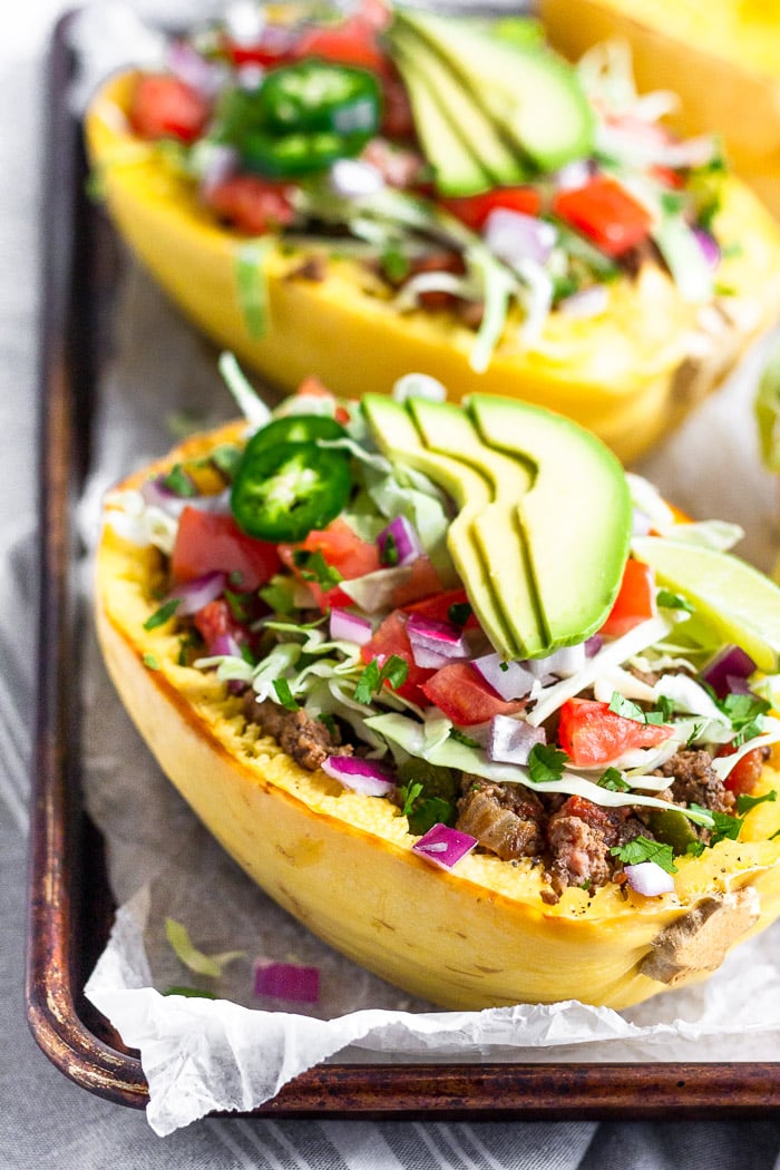 Two taco spaghetti squash boats on a baking tray topped with toppings and garnished with a lime wedge.