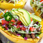 A baking tray with taco stuffed spaghetti squash boats on top that are topped with cabbage, diced tomatoes, avocado, cilantro, and jalapeños.