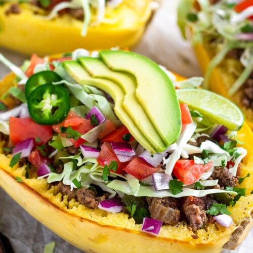 A baking tray with taco stuffed spaghetti squash boats on top that are topped with cabbage, diced tomatoes, avocado, cilantro, and jalapeños.