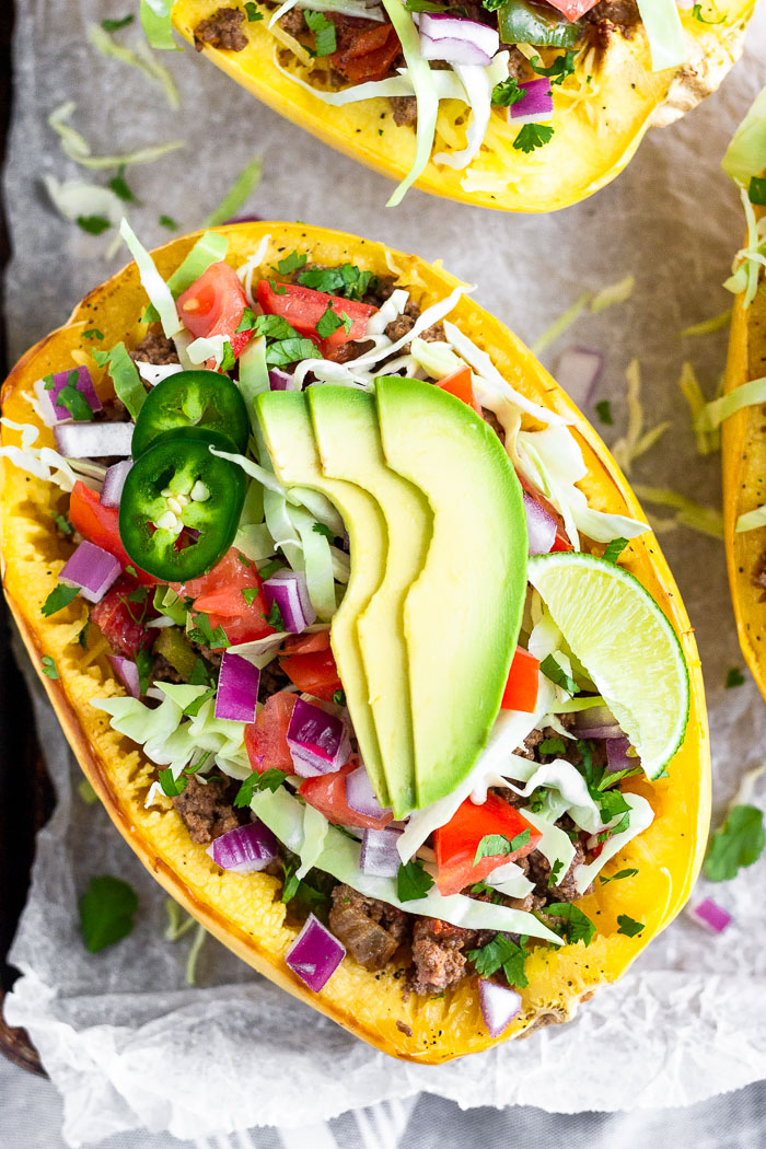 Overhead shot of a taco stuffed spaghetti squash boat topped with shredded cabbage, diced tomato, diced red onion, sliced jalapeños, sliced avocado, and a lime wedge.