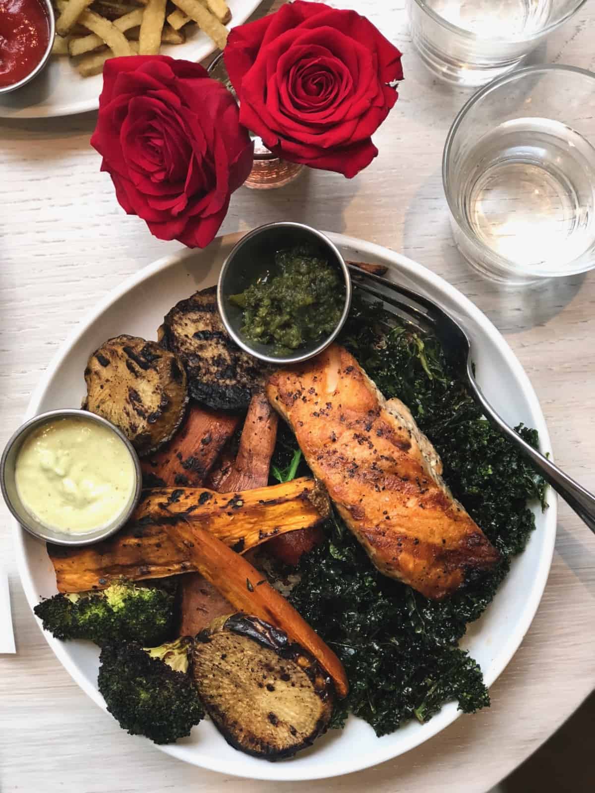 A table of a bowl of crispy kale, roasted veggies, and salmon with two sauces on the side, two red roses, and a glass of water