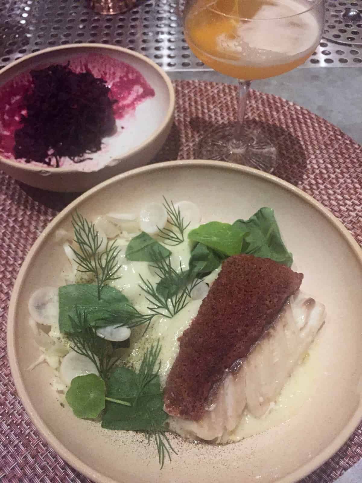 Piece of fish with greens and a bowl of beets and a cocktail in the background