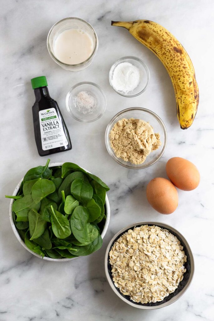 Marble counter top with a glass of milk, one ripe banana, a bowl of baking powder, a bowl of protein powder, two brown eggs, a bowl of oats, a bowl of spinach, a bottle of vanilla extract, and a bowl of salt.