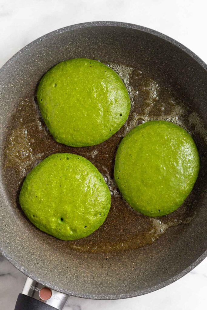 Green pancakes cooking on a skillet before they are flipped.