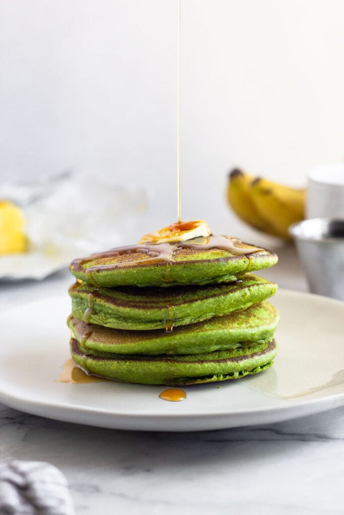 A stack of spinach banana pancakes topped with butter and maple syrup being poured on them. Behind them is a stick of butter, some ripe bananas, and a small ramekin of maple syrup.
