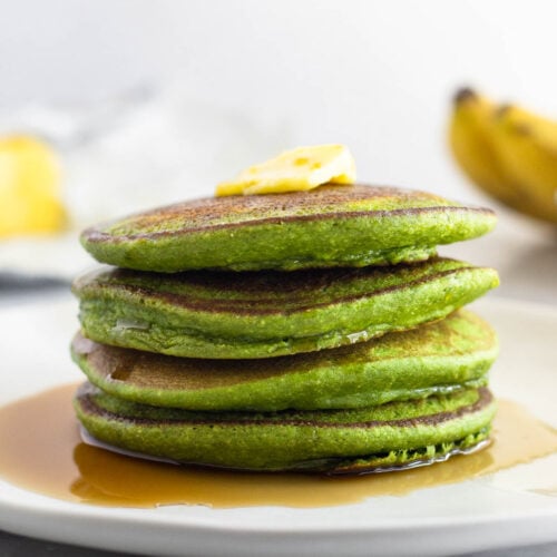 A plate of four spinach pancakes stacked on top of each other with butter and syrup on them. Behind them are some ripe bananas and more butter.