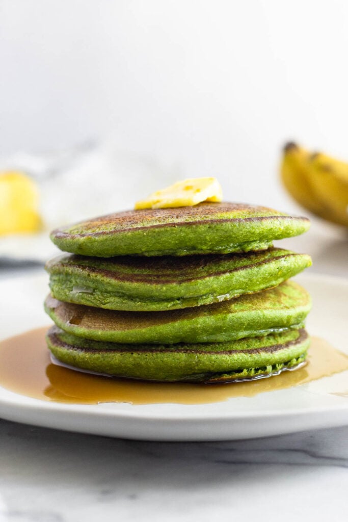 A plate of four spinach pancakes stacked on top of each other with butter and syrup on them. Behind them are some ripe bananas and more butter.