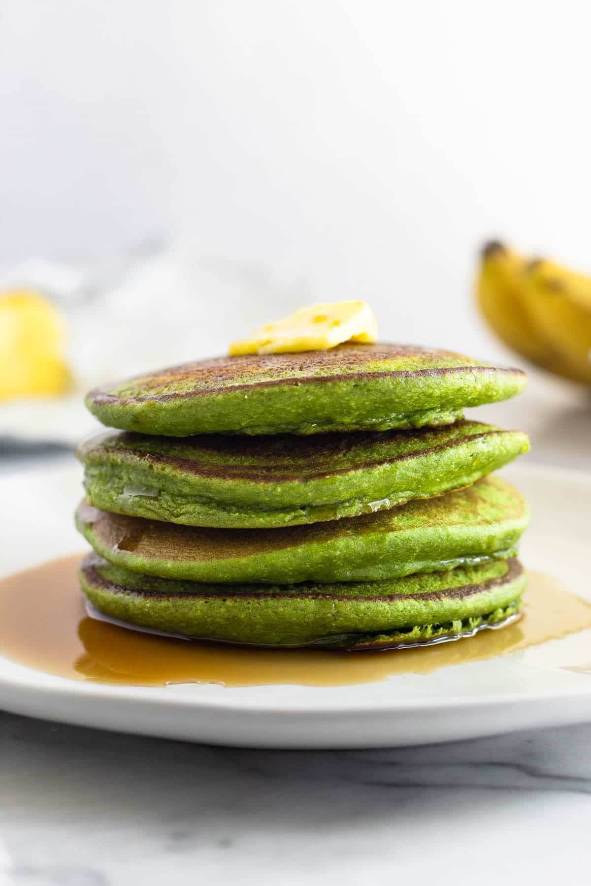 A plate of four spinach pancakes stacked on top of each other with butter and syrup on them. Behind them are some ripe bananas and more butter.