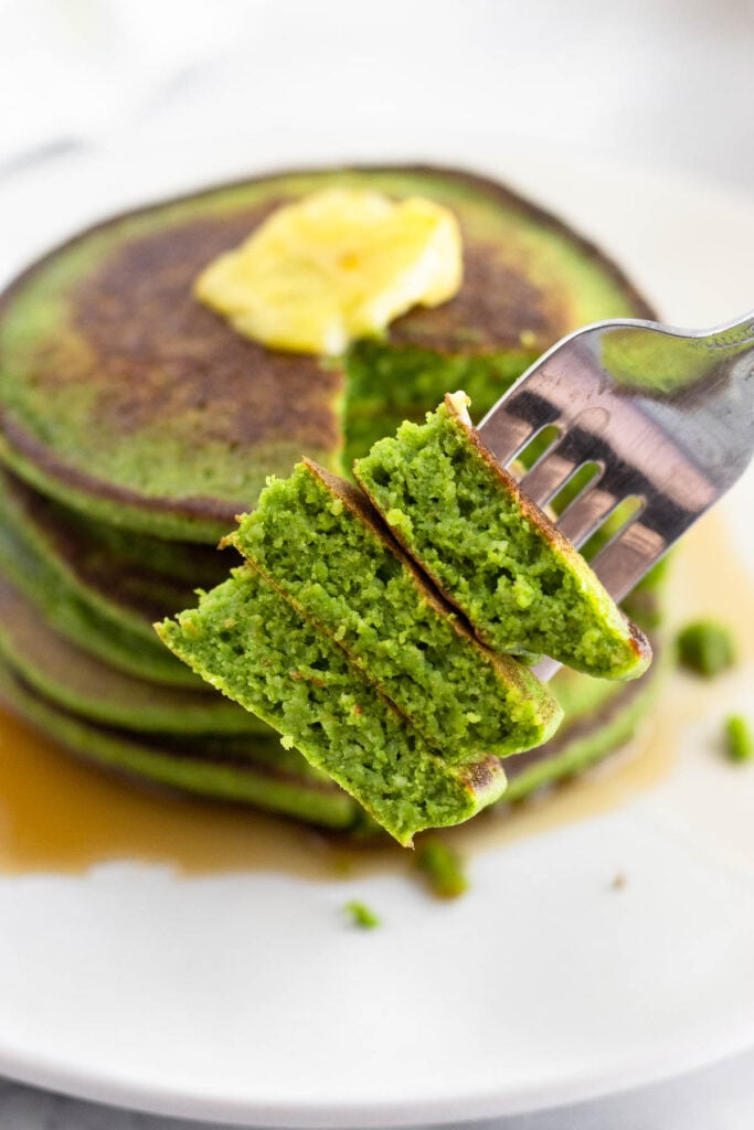 Spinach pancakes stacked on a plate with butter on them. There ia fork in the forefront with some on it showing the inside.