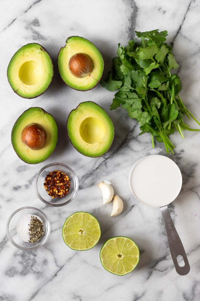 White marble countertop with a bunch of cilantro on it, a half cup of coconut milk, a lime cut in half, a bowl of salt and pepper, a bowl of red pepper flakes, and 4 halves of avocados.