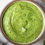 Creamy avocado sauce in a bowl. Around it is a bowl of tortilla chips, some cilantro, a lime wedge, and a striped kitchen towel.