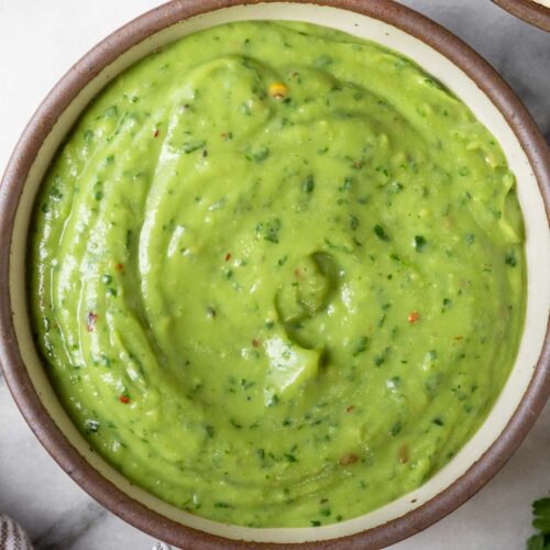 Creamy avocado sauce in a bowl. Around it is a bowl of tortilla chips, some cilantro, a lime wedge, and a striped kitchen towel.