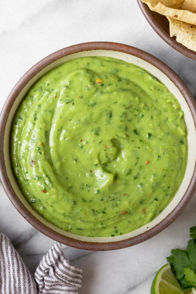 Creamy avocado sauce in a bowl. Around it is a bowl of tortilla chips, some cilantro, a lime wedge, and a striped kitchen towel.
