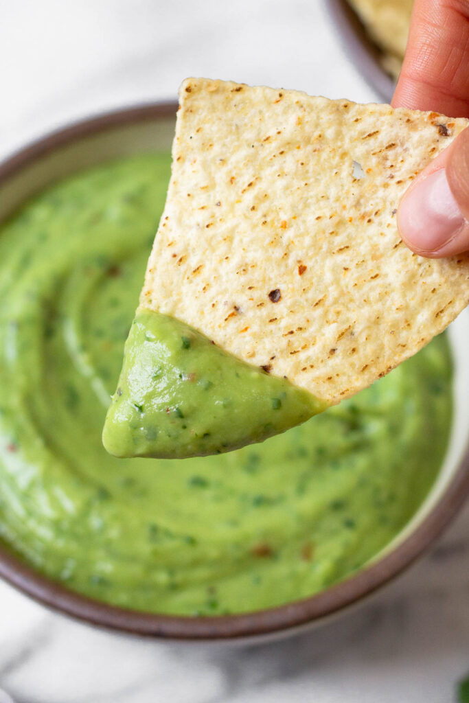 A hand holding a chip with avocado lime sauce on it. Behind it is a bowl of more sauce.