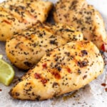 A plate filled with grilled lemon pepper chicken with lemon wedges. Half a lemon is off in the distance behind the plate.