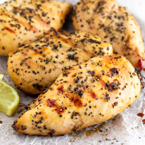 A plate filled with grilled lemon pepper chicken with lemon wedges. Half a lemon is off in the distance behind the plate.