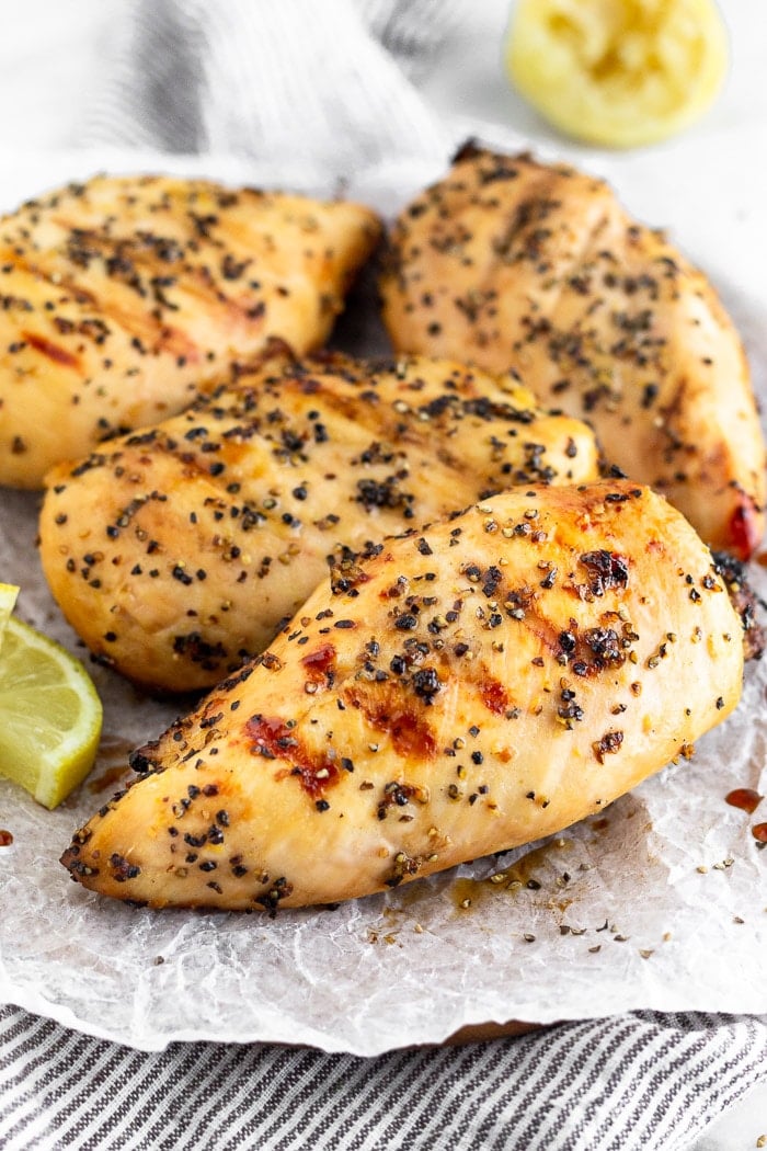 A plate filled with grilled lemon pepper chicken with lemon wedges. Half a lemon is off in the distance behind the plate.