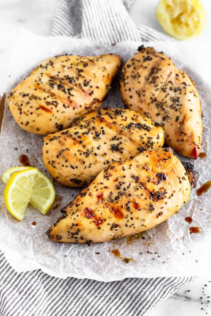 Large plate with parchment paper filled with grilled lemon pepper chicken with lemon wedge on the side. Behind it is a lemon wedge and there is pepper off the side of the plate.