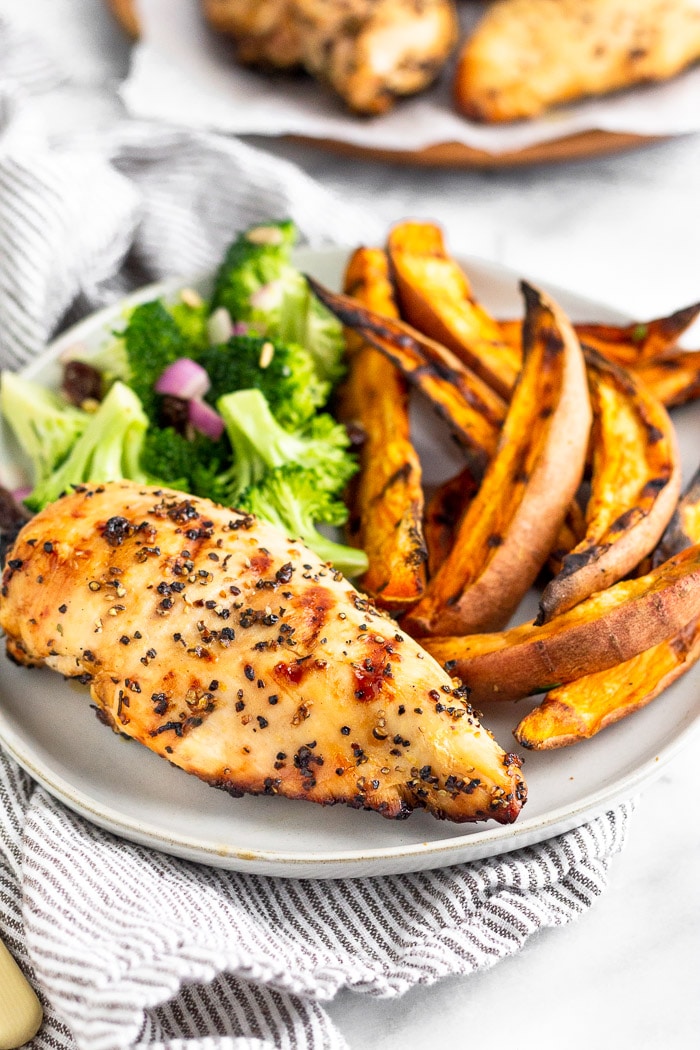 A plate filled with grilled chicken. sweet potato fries, and broccoli salad. It is sitting on a stripped towel with a large plate of chicken behind it.