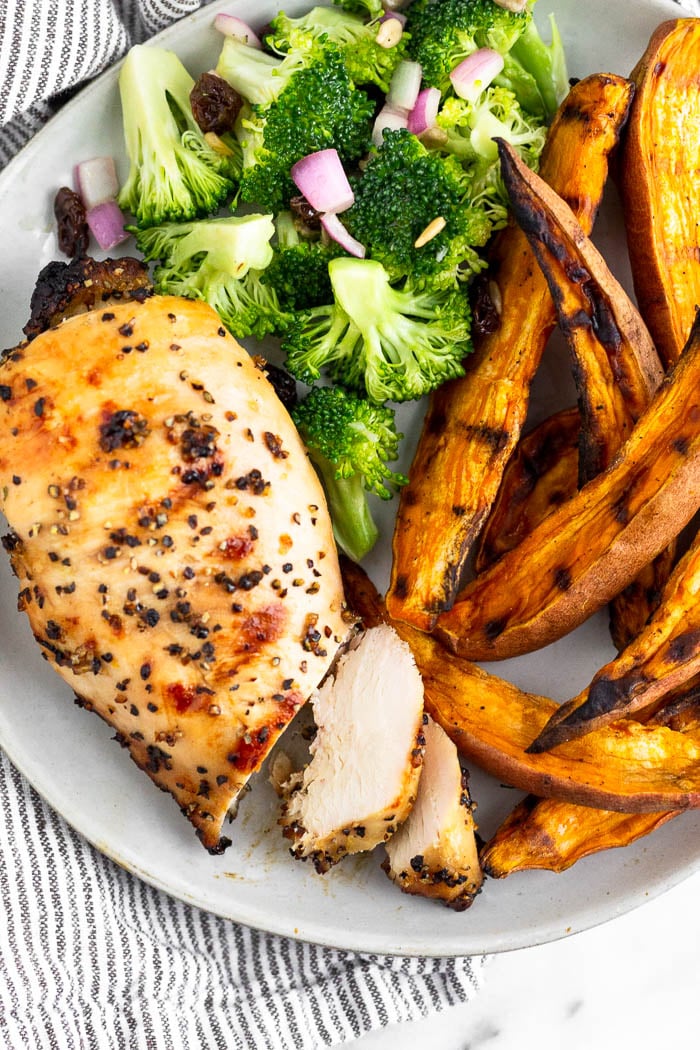 Plate filled with a piece of grilled lemon chicken that has 2 pieces cut from it. Next to it are grilled sweet potato fries and a broccoli salad.