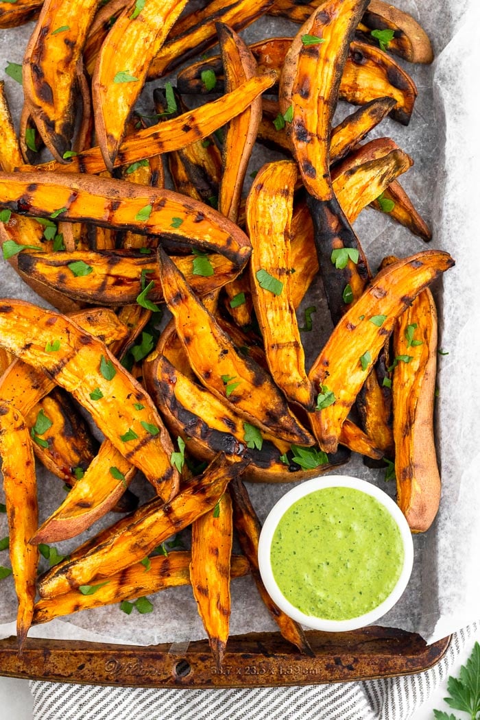 Baking sheet with grilled sweet potato fries topped with fresh parsley an a green dipping sauce.