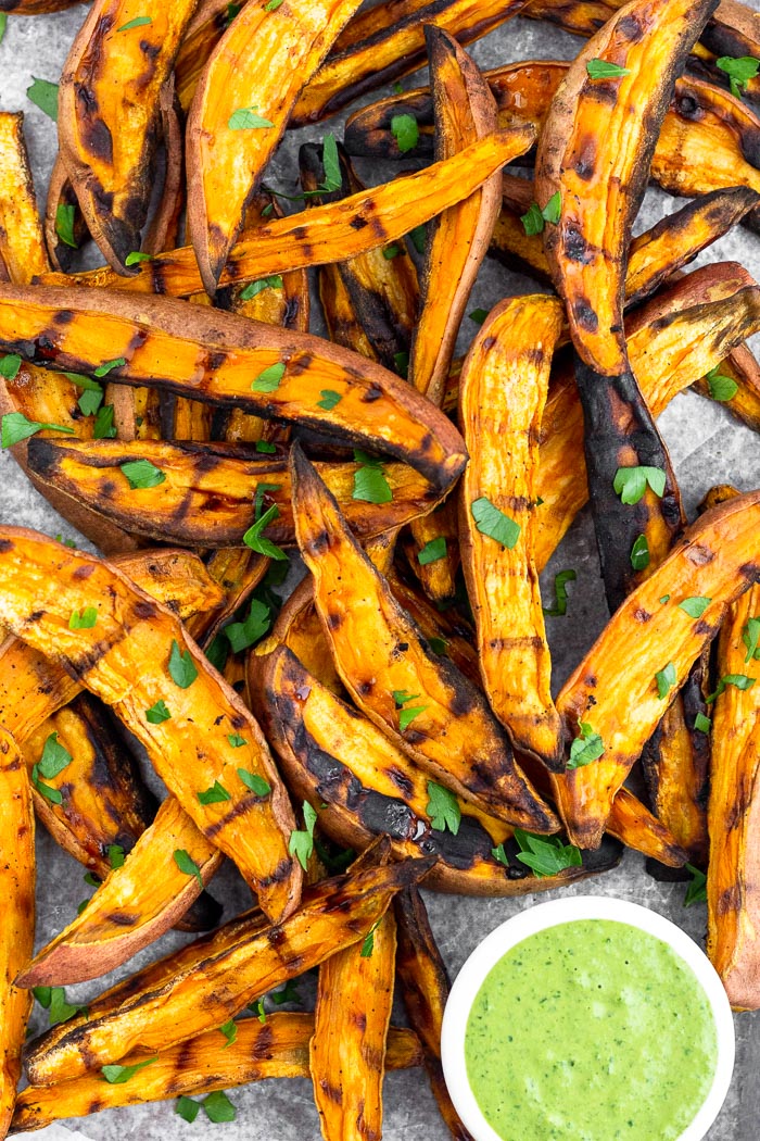 Close up of a bunch of grilled sweet potato wedge fries topped with fresh chopped parley and a green tahini dipping sauce in a small saucer next to them.