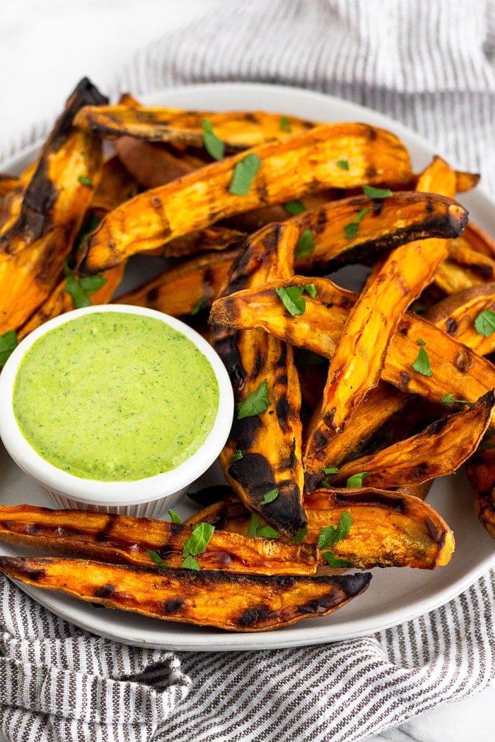 Small white plate filled with sweet potato fries made on the grill. They are topped with fresh chopped parsley and surrounding a parsley tahini green dipping sauce.
