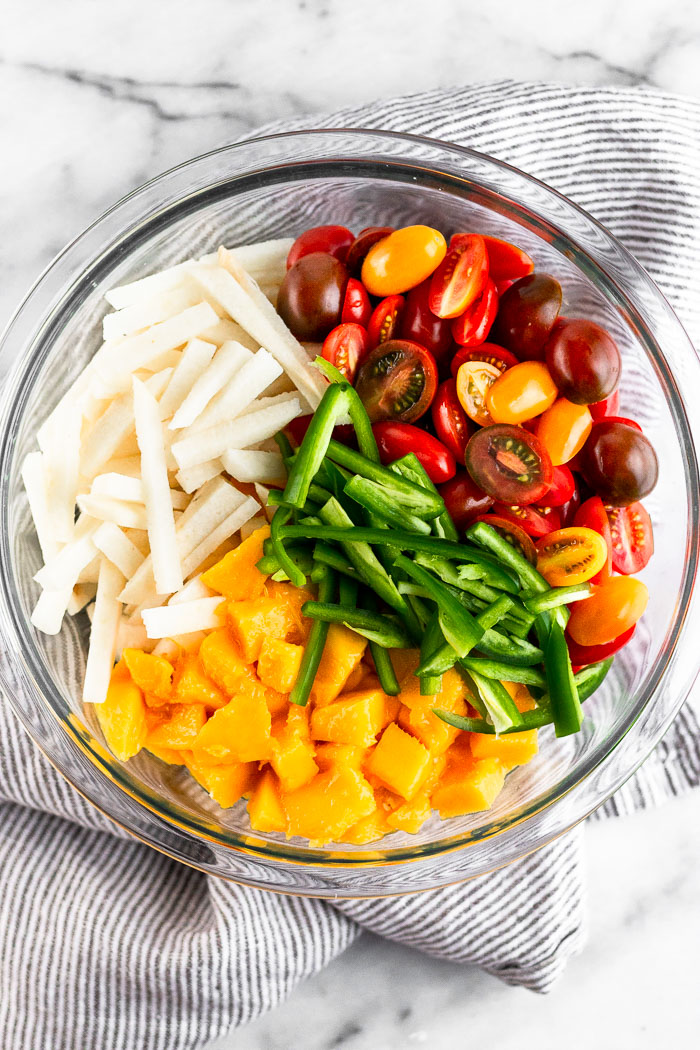 Bowl filled with sliced jicama, halved cherry tomatoes, sliced jalapeños, and diced mango. The bowl is sitting on a stripped towel.