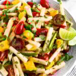 Bowl filled with jicama salad with tomatoes, mango, and jalapeños garnished with lime wedges. It is sitting on a stripped towel with half a lime and cilantro behind it.