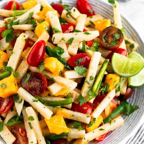 Bowl filled with jicama salad with tomatoes, mango, and jalapeños garnished with lime wedges. It is sitting on a stripped towel with half a lime and cilantro behind it.