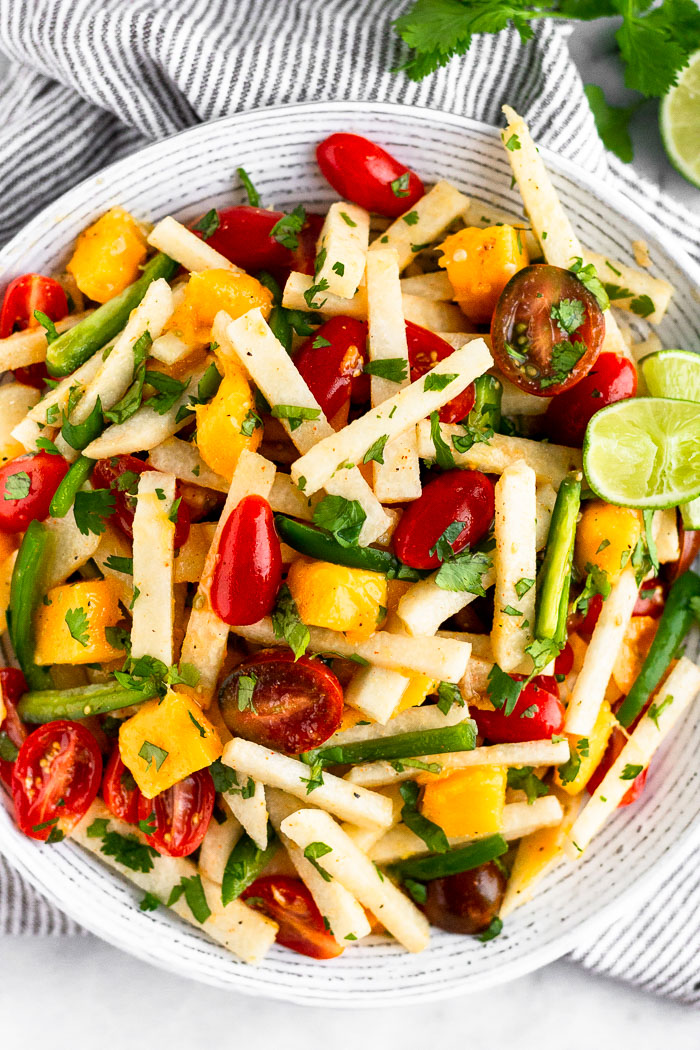 Overhead shot of jicama salad with tomatoes, mango, and jalapeños. Lime wedges are garnishing the plate that is on top of a stripped towel.