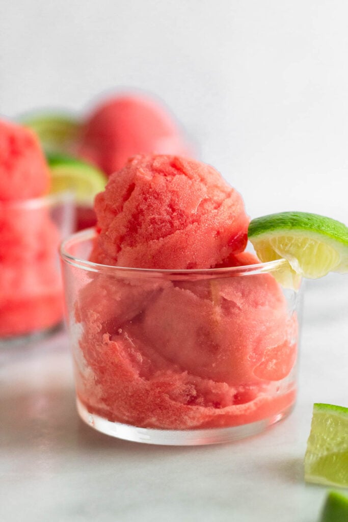 Watermelon lime sorbet in a small glass with a lime wedge. Next to it is a lime wedge and behind it is two more glasses.