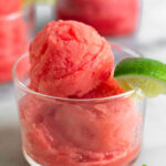 Watermelon sorbet in a small glass with a lime wedge. Behind it is two more glasses.
