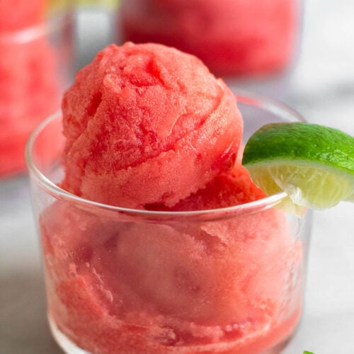 Watermelon sorbet in a small glass with a lime wedge. Behind it is two more glasses.