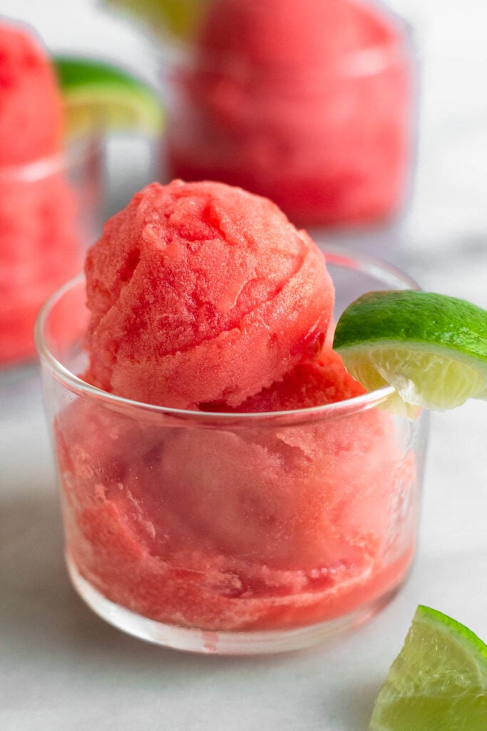 Watermelon sorbet in a small glass with a lime wedge. Behind it is two more glasses.