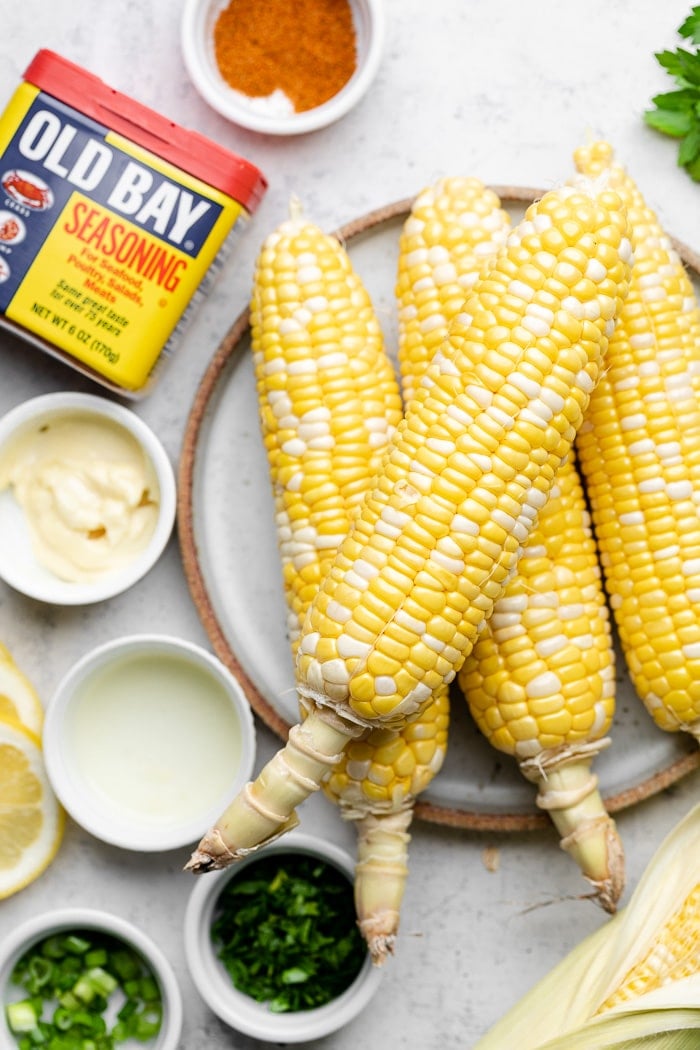 Overhead shot of husked corn on the cob, small bowls of herbs, bowl of mayo, and and Old Bay container with it in a bowl next to it.